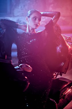 Siyah uzun gece elbisesi giymiş güzel bir motosikletli kız garajda motosiklet üstünde poz veriyor..