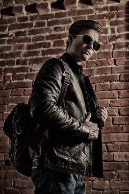 Yakışıklı genç adam, sırtında sırt çantasıyla güneş gözlüğü ve deri ceketi ile şehirde bir duvarın yanında poz veriyor. Yaşam tarzı ve erkek modası. 