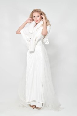 Güzellik, moda. Pahalı beyaz bir gece elbisesi ve beyaz vizon peleriniyle poz veren, sarı kıvırcık saçlı, alımlı bir kadın. Lüks bir şekilde. Gelinlik. Tam boy çekim.