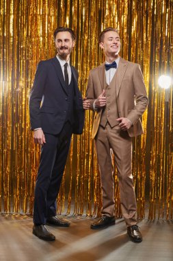 Noel ve yeni yıl modası. Şık takım elbiseli iki yakışıklı genç adam parlak altın perdelerin arkasında duruyor ve gülüyorlar. Erkek modası.