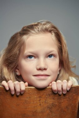 Gülen, gülümseyen, 11 yaşında, dalgalı sarı saçlı, yüzü ve elleri sandalyeye yaslanmış bir kızın yakın portresi. Gri stüdyo geçmişi. Mutlu çocukluk. 