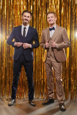 Noel ve yeni yıl modası. Zarif gece kıyafetleri içindeki iki yakışıklı genç adam parlak altın folyo perdelerin arkasında neşeli bir şekilde gülümsüyor. Erkek modası.