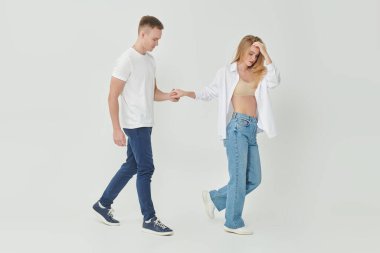 Stüdyoda beyaz gömlekli ve kot pantolonlu güzel bir çiftin beyaz arka planda yürürken çekilmiş bir fotoğrafı. Aşk ve moda. Tam boy portre. 