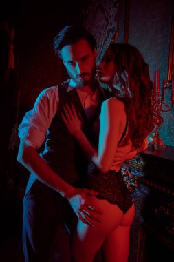 Aşık göz kamaştırıcı bir çift. Seksi siyah iç çamaşırlı güzel bir kadın ve şık takım elbiseli yakışıklı bir adam, koyu lüks, kırmızı ışıklı bir odada birbirlerine tutkuyla sarılıyorlar. Moda fotoğrafı.. 