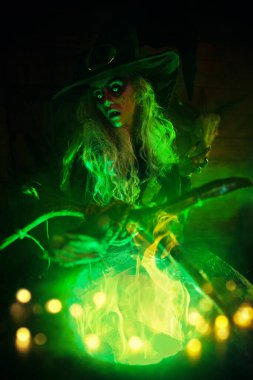 Korkunç yaşlı cadı, sihirli yeşil ışıkla çevrili kazanında büyücülük yapıyor. Cadılar Bayramı. Korkunç hikayeler..