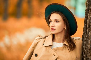 Ağacın yanındaki sonbahar parkında beyaz yün kazak, ceket ve şapka giymiş zarif genç bir bayan poz veriyor. Sonbahar modası ve güzelliği. 