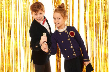 Zarif gece kıyafetleri içinde neşeli çocuklar arka planda parlak altın folyo perdelerle mikrofona şarkı söylüyorlar. Çocuk partisi. Çocuk modası ve tatiller. 