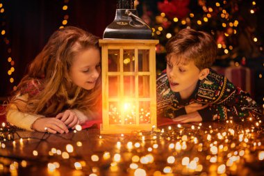 Mutlu küçük kız ve oğlan bir Noel ağacının yanında uzanır ve güzel bir fener ve ışığa hayran kalırlar. Noel ve Mutlu Yıllar Kutlaması. 