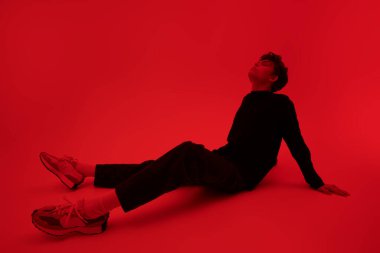 Kapalı gözlü ve yüzünde üzgün bir ifadeyle parlak kırmızı bir arka planda oturan yakışıklı bir adamın tam boy portresi. Gençlik sorunları ve duyguları.