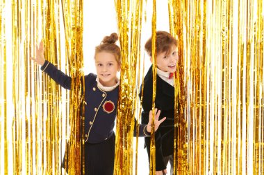 Zarif gece kıyafetleri içinde iki neşeli çocuk parlak altın folyo perdelerin arkasından neşeyle gülümsüyor. Çocuk partisi. Çocuk modası ve tatiller. 