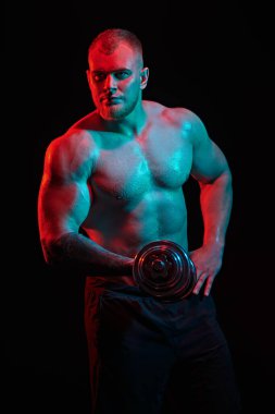 Güçlü, sağlıklı, atletik bir adamın portresi. Kırmızı ışıklı siyah bir arka planda dambıllarla sıkı çalışıyor. Erkek güzellik konsepti. Fitness, vücut geliştirme.