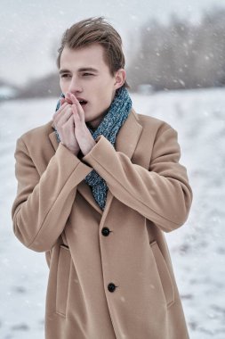 Soğuk ve karlı bir günde, şık ceketli ve eşarplı yakışıklı genç bir adam donmuş ellerini ısıtıyor. Moda fotoğrafı. İnsan konsepti, yaşam tarzı.