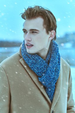 Paltosu ve atkısı olan yakışıklı sarışın genç adam soğuk ve karlı bir günde dışarıda duruyor. Moda fotoğrafı. İnsan konsepti, yaşam tarzı.