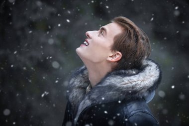 Tarz sahibi siyah deri ceketli, kürklü, parkta kar yağışının keyfini çıkaran sarışın bir adamın yan görüntüsü. Erkek modası. Taze kış günü.