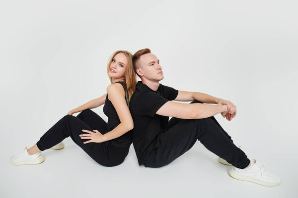 Happy young loving couple in black sportswear and white sneakers sitting back-to-back on a white background. Love and fashion.