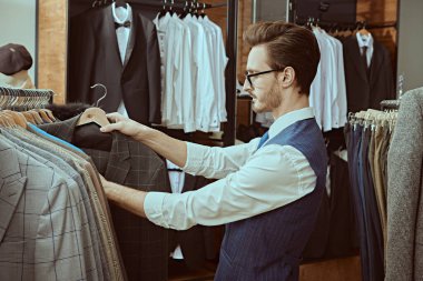 Zarif klasik takım elbiseli ve gözlüklü yakışıklı bir esmer premium erkek giyim mağazasından yeni bir takım elbise seçiyor. Erkek işi modası. Yan plan çekimi.