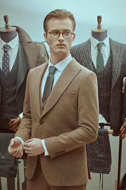 Zarif kahverengi takım elbiseli ve gözlüklü yakışıklı sarışın bir adam lüks klasik takım elbiselerin arka planında ciddi ciddi başka tarafa bakıyor. Erkeklerin iş modası.