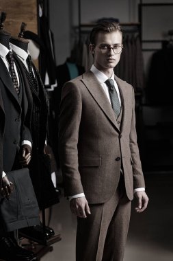 Portrait of a serious handsome blond man in an elegant brown three-piece suit and glasses standing in a premium men's clothing store in dim lighting. Men's business fashion. clipart