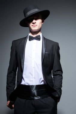 Siyah smokinli yakışıklı bir genç. Papyonlu ve şapkalı. Gri stüdyo arka planında gölgesi var. Erkek modası ve güzelliği. 