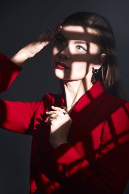 Gece makyajlı güzel genç bir kadın yüzünde gölgeler olan koyu gri bir arka planda zarif kırmızı bir takım elbiseyle poz veriyor. Kadınlık, feminizm ve moda. Zarif bir iş tarzı.. 