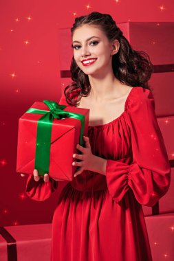 Kırmızı elbiseli, gece makyajlı ve saç stili güzel, neşeli kız bir hediye taşıyor. Kırmızı arka planda bir sürü kırmızı hediye kutusu ve etrafta uçuşan sihirli ışıklar var. Noel ve Yeni Yıl Kutlaması. 