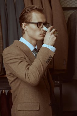 Zarif kahverengi takım elbiseli yakışıklı sarışın ve gözlüklü bir adam lüks klasik takım elbisesinin arka planında viski içiyor. Erkek işyeri modası ve yaşam tarzı.