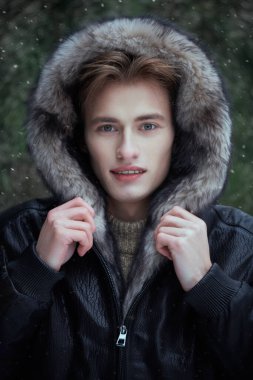 Çekici, siyah deri ceketli, başında kürk kapüşonlu, kar yağışlı bir günde köknar ağaçlarının arka planında gülümseyen çekici sarışın bir adam. Erkekler kış modası.