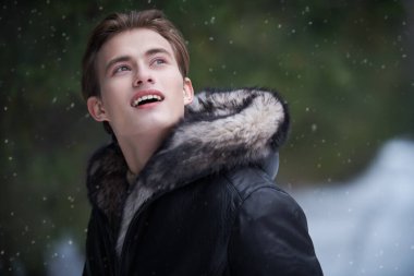 Parkta kürklü siyah deri ceketli genç sarışın adam duruyor ve karlı bir kış gününde doğanın güzelliğine hayran. Erkek modası. Kış mevsimi. 