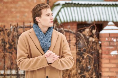 Soğuk bir kış gününde tuğladan bir evin yanında duran şık ceketli ve eşarplı yakışıklı bir gencin portresi. Donmuş ellerini ısıtıyor ve bir şeyler düşünüyor. Moda ve yaşam tarzı.