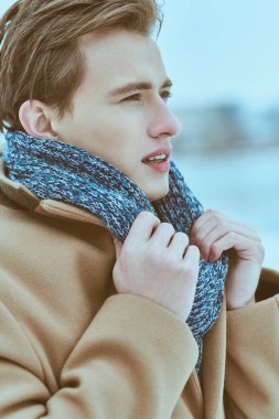 Şık ceketli ve atkılı yakışıklı sarışın genç adam soğuk bir kış gününde dışarıda duruyor. Kış erkek modası. İnsan konsepti, yaşam tarzı.