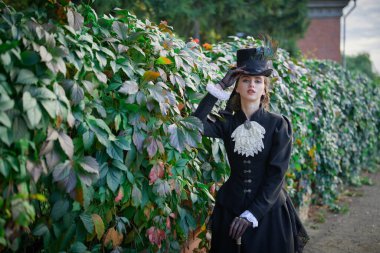 Katı siyah elbiseli ve şapkalı zarif esmer kadın arka planda poz veriyor. 19. ve 20. yüzyılın başları. Tarihsel makyaj ve saç stili.