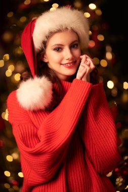 Kırmızı kazaklı ve Noel Baba şapkalı güzel bir kız Noel ağacının ve yanıp sönen ışıkların arkasına gülümsüyor. Noel ve Yeni Yıl Kutlaması.