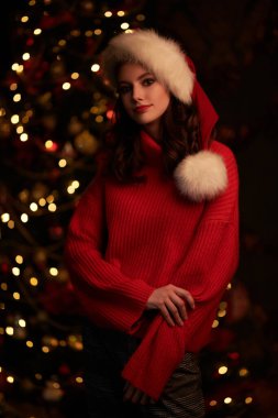 Noel ve Yeni Yıl kutlaması. Kırmızı kazaklı ve Noel Baba şapkalı güzel bir kız Noel ağacının karanlık arka planına ve yanıp sönen ışıklara gülümsüyor..