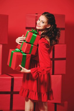 Güzel kırmızı bir elbise giyen, akşam makyajlı ve saç stili olan mutlu kız bir sürü hediye taşıyor. Kırmızı arka planda bir sürü kırmızı hediye kutusu var. Noel ve Yeni Yıl indirimi. 