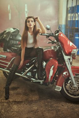 Diz kapağında siyah deri, deri şort ve üstü eski bir garajda motosikletinin üzerinde oturan kendine güvenen güzel bir motorcu kız. Tam boy portre.
