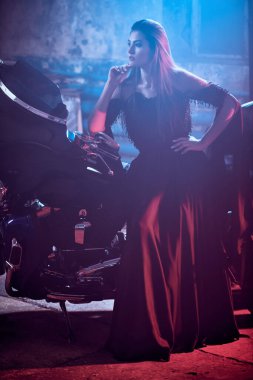 Uzun siyah bir gece elbisesi içinde zarif makyajlı, göz kamaştırıcı genç bir kadının moda çekimi garajda kırmızı ve mavi ışıkla aydınlatılmış bir motosikletin yanında poz veriyor. Tam boy portre.