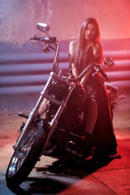 Zarif makyajlı ve siyah bir gece elbisesi içinde uzun düz saçlı güzel bir kız garajda kırmızı ışıkla aydınlatılmış bir motosiklette poz veriyor. Tam boy portre. Moda fotoğrafı.. 