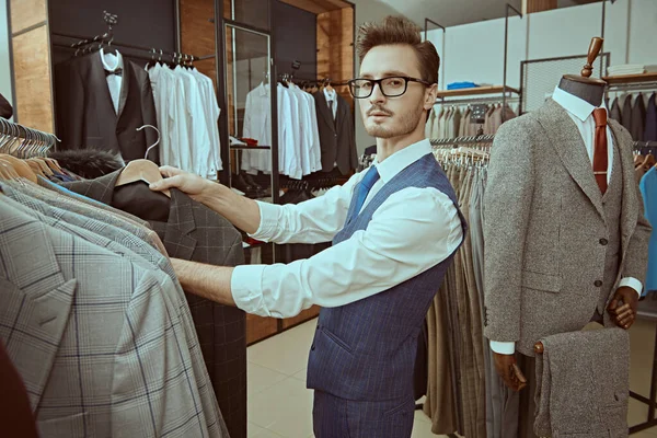 A handsome brunet man in elegant classic suit and glasses chooses a new suit in a premium men\'s clothing store. Men\'s business fashion.