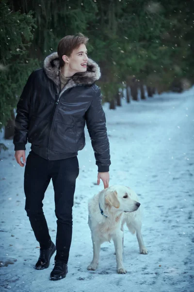 Şık siyah deri ceketli neşeli sarışın adam köpeğiyle kış parkında yürüyor. Erkek modası. Mutlu sahibi ve evcil köpeği. Tam boy çekim.
