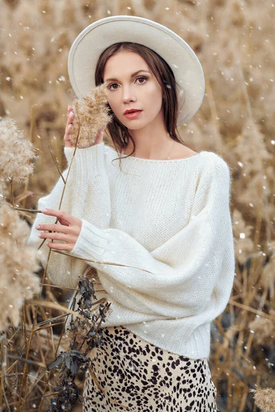 Beyaz yün kazak ve beyaz keçeli şapkalı zarif genç kadın sonbahar parkında sazlıklar ve düşen kar taneleri arasında poz veriyor. Kış modası ve güzellik. 