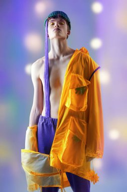 Gençlik tarzı ve yaratıcı tasarımcı kıyafetleri. Parlak sarı ceketli, mor eşofmanlı, başında yeşil şapkalı, açık mor bir arka planda gözleri kapalı poz veren havalı bir adam.. 