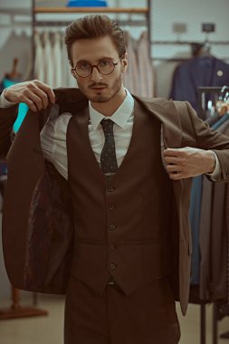 Gözlüklü, zengin, yakışıklı bir esmer adamın portresi ve bir erkek giyim mağazasında kameraya bakan üç parçalı şık bir takım elbise. Erkek modası, iş tarzı.. 