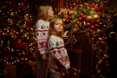 Mutlu Noeller ve yeni yıl. Örgü kazaklı iki küçük kız kardeş karanlık bir odada Noel ağacı ve ışıklar yanıp sönerken güzel süslemelere bakarlar..