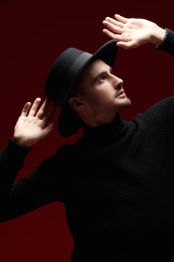 Siyah balıkçı yaka kazaklı yakışıklı esmer adam ve kırmızı stüdyo arka planında profil pozu veren bir şapka. Erkek güzelliği ve tarzı. 