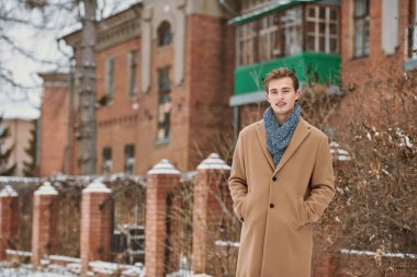 Şehir caddesinde tuğladan bir evin yanında duran şık montlu ve atkılı neşeli yakışıklı genç adam. Erkek modası ve yaşam tarzı.