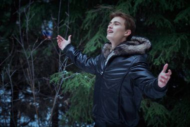 Tüylü kapüşonlu siyah deri ceketli sarışın genç adam kış parkında kollarını açıp doğanın güzelliğine hayran kalıyor. Erkek modası. Harika kış mevsimi. 