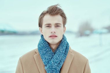 Şık ceketli ve atkılı yakışıklı sarışın genç adam soğuk bir kış gününde dışarıda duruyor. Moda fotoğrafı. İnsan konsepti, yaşam tarzı.
