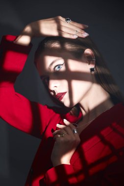 Gece makyajlı güzel genç bir kadın yüzünde gölgeler olan koyu gri bir arka planda zarif kırmızı bir takım elbiseyle poz veriyor. Kadınlık, feminizm ve moda. Zarif bir iş tarzı.. 