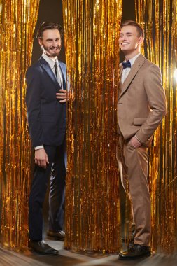 Noel ve yeni yıl modası. Şık takım elbiseli iki yakışıklı genç adam parlak altın folyo perdelerin arkasında neşeli bir şekilde gülümsüyor. Erkek modası.