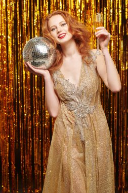 Şampanyası ve parlayan disko topuyla lüks altın bir elbise içinde poz veren güzel kızıl saçlı kadın. Parlak altın folyo perdeler arka planda. Parti ve kutlama. Tatil modası.  
